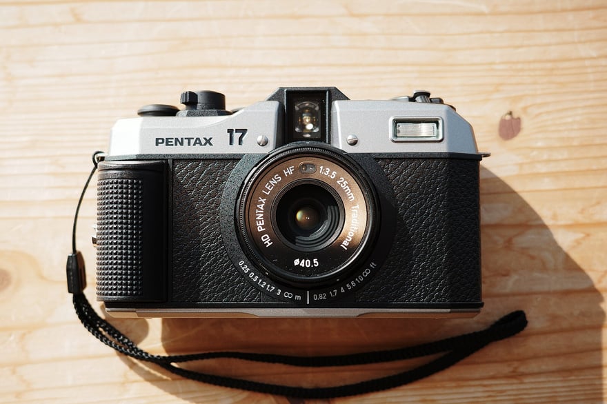 Pentax 17 की सामने से फ़ोटो: चाँदी जैसे ऊपरी कवर और काली चमड़े-जैसी बनावट वाली बॉडी का हाफ़-फ़्रेम फ़िल्म कैमरा, लेंस रिंग पर HD Pentax Lens HF 1:3.5 25mm लिखा है, लकड़ी की मेज़ पर रखा है, काली कलाई-पट्टी लगी है।