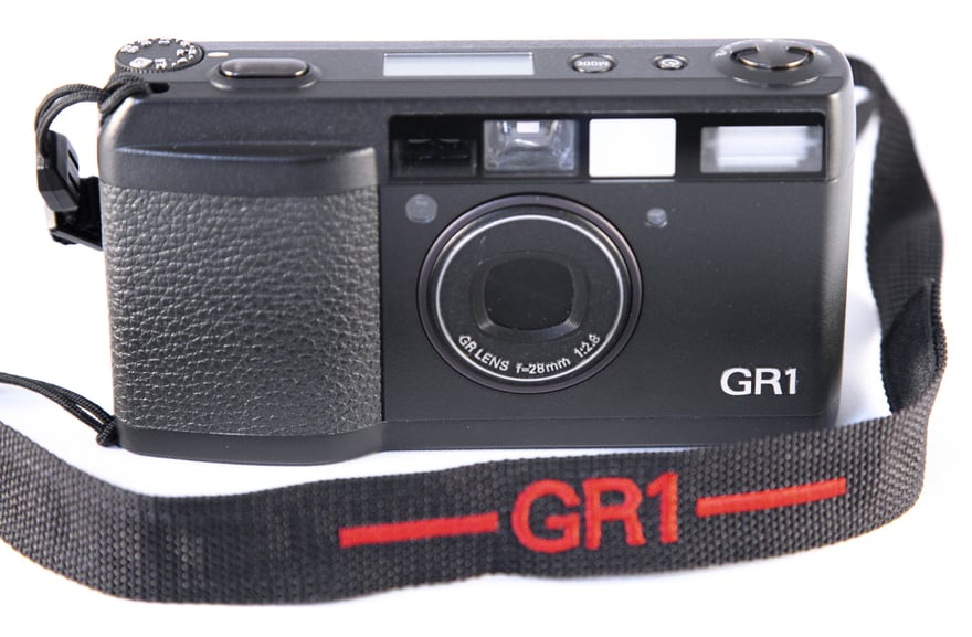 Ricoh GR1 की सामने से फ़ोटो: काली बॉडी वाला छोटा फ़िल्म कैमरा, लेंस रिंग पर GR LENS 1:2.8 28mm लिखा है, बॉडी के नीचे दाईं ओर GR1 छपा है, आगे लाल GR1 अक्षरों वाला काला स्ट्रैप लटक रहा है।