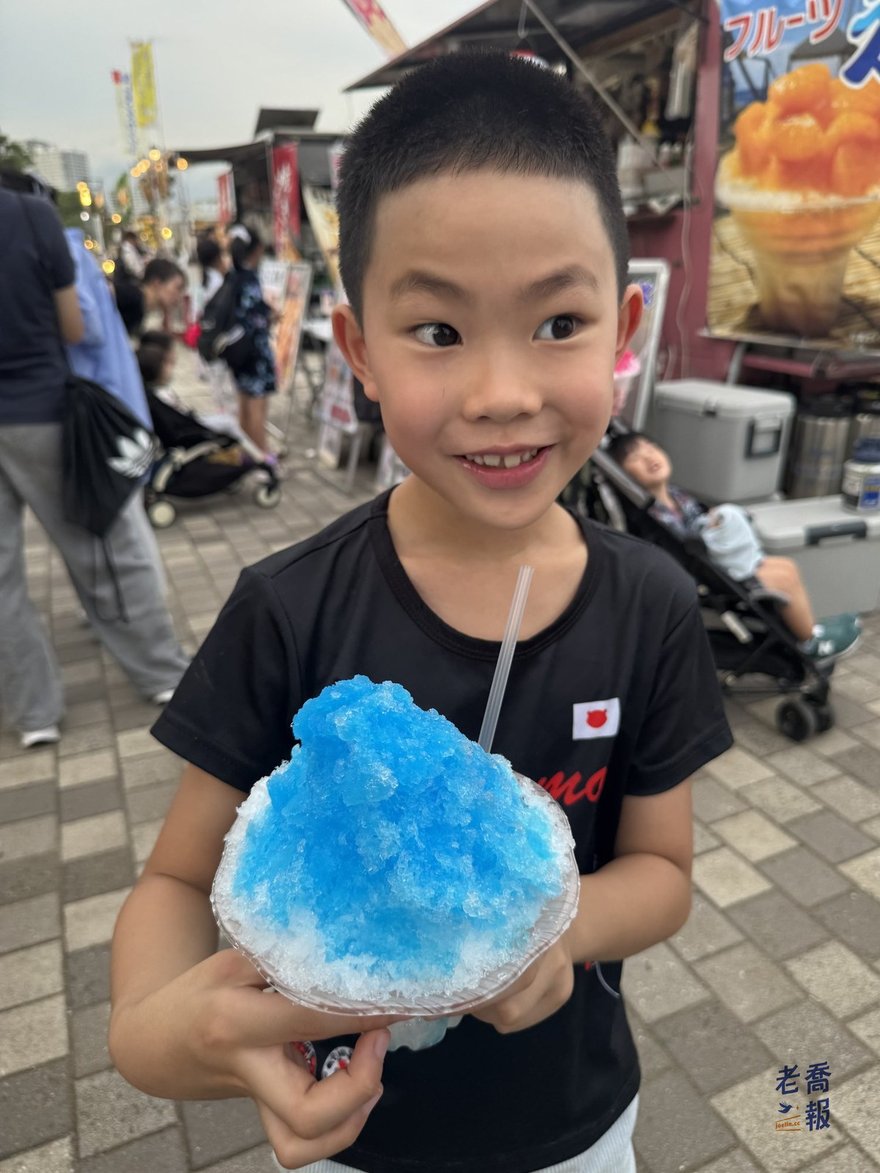 嘻嘻雙手捧著堆得很高的藍色刨冰，背景是祭典攤位與燈串