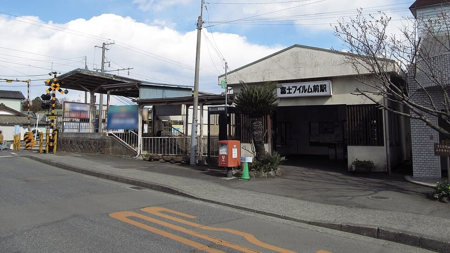 伊豆箱根鐵道大雄山線的富士フイルム前站，一棟灰色平房站舍，招牌寫著「富士フイルム前駅」，左側是平交道與月台遮棚。