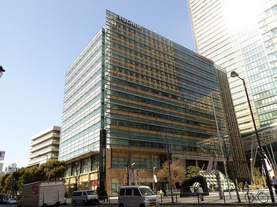 2015 年的東京中城富士軟片總部大樓，頂端掛著 FUJIFILM 字樣，右側側面同時掛著紅色的 FUJI XEROX 招牌，一樓入口處也有 FUJIFILM 字樣，前景是街道與車輛。