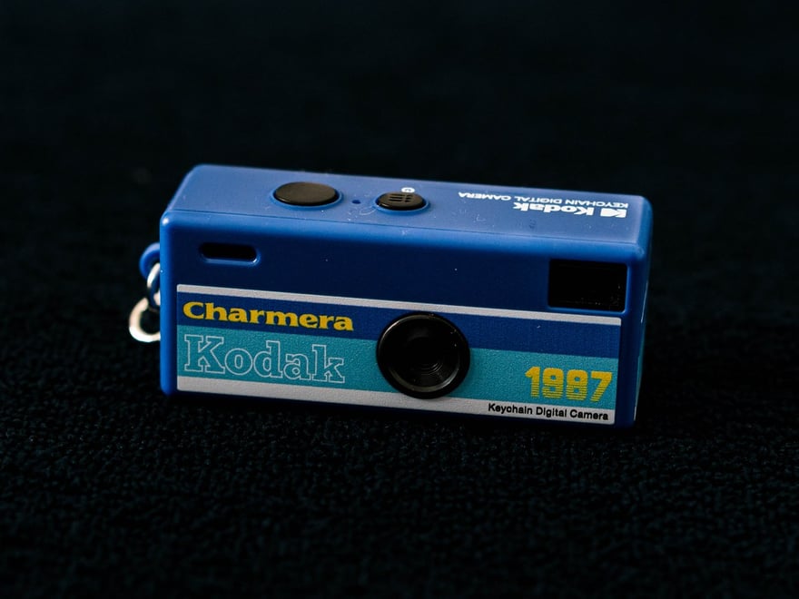 Kodak Charmera 鑰匙圈相機的正面特寫：藍色長方形小機身，左側有金屬鑰匙圈扣環，機身下半部是淺藍與白色的橫條，印著黃色 Charmera、描邊的 Kodak 字樣、1987 字樣與 Keychain Digital Camera，中間是小小的黑色鏡頭，背景是黑色絨布。