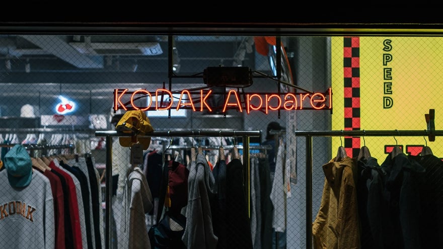 東京原宿 Kodak Apparel 門市的櫥窗：玻璃後方懸著橘紅色霓虹燈招牌「KODAK Apparel」，下方衣架掛著外套、帽子與印有 KODAK 字樣的大學 T，右側是黃底紅黑格紋的牆面。這家店由韓國授權商經營，不是 Kodak 開的。