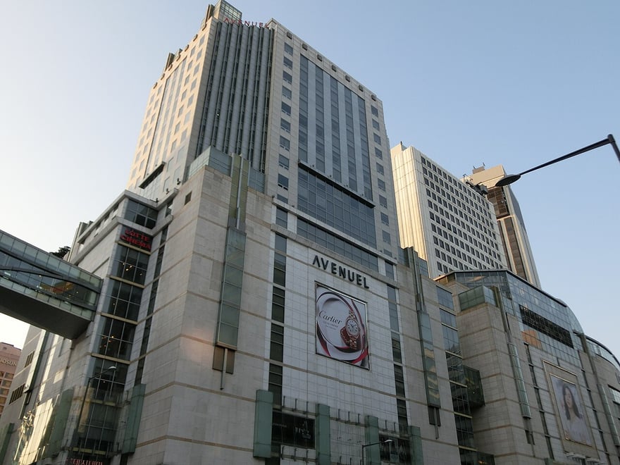 首爾明洞的樂天百貨總店：灰白色的大樓外牆，中段掛著 AVENUEL 的招牌與大幅廣告看板，旁邊有連到隔壁大樓的空橋。