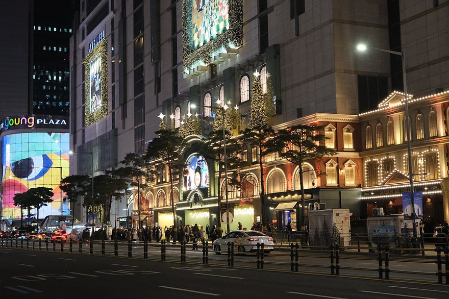 首爾明洞樂天免稅店一帶的夜景：大樓外牆的巨型 LED 螢幕亮著，街道兩旁的行道樹掛滿燈飾。
