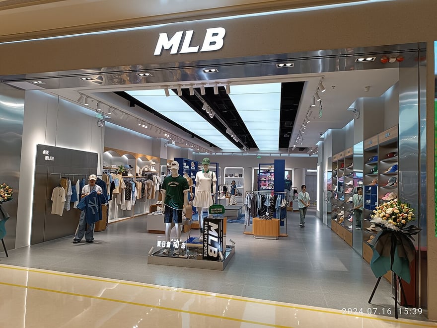 中國深圳寶安海岸城購物中心的 MLB 服飾店：白色門面上方是銀色 MLB 字樣，店內貨架陳列棒球帽、上衣與球鞋，入口擺著開幕花籃。