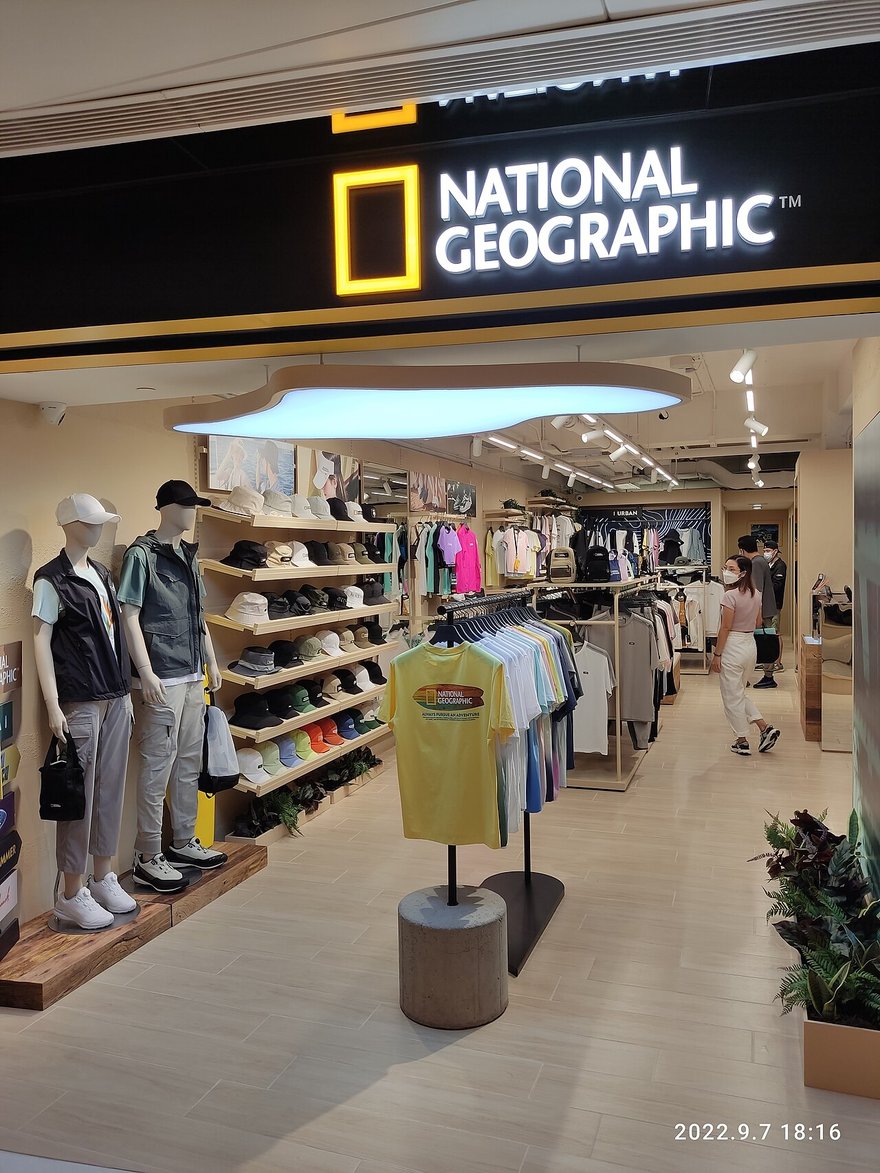香港沙田新城市廣場的國家地理服飾店：黑色門楣上是黃框國家地理標誌，店內陳列帽子、上衣、外套與鞋子，門口有兩個穿著背心的假人。