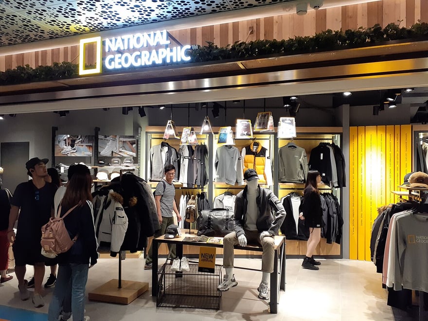 香港尖沙咀 K11 MUSEA 購物中心的國家地理服飾店：門楣上方是黃框國家地理標誌，店內木質層架陳列外套、背包與上衣，幾位顧客在挑選。