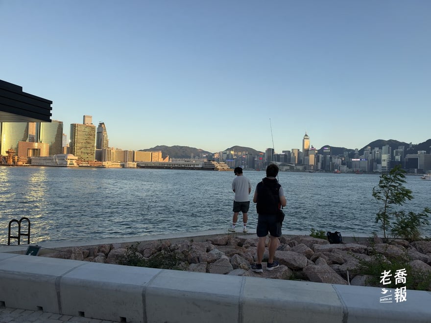西九海濱的石堤上，兩個男生背對鏡頭站著釣魚，對岸是中環的高樓和太平山