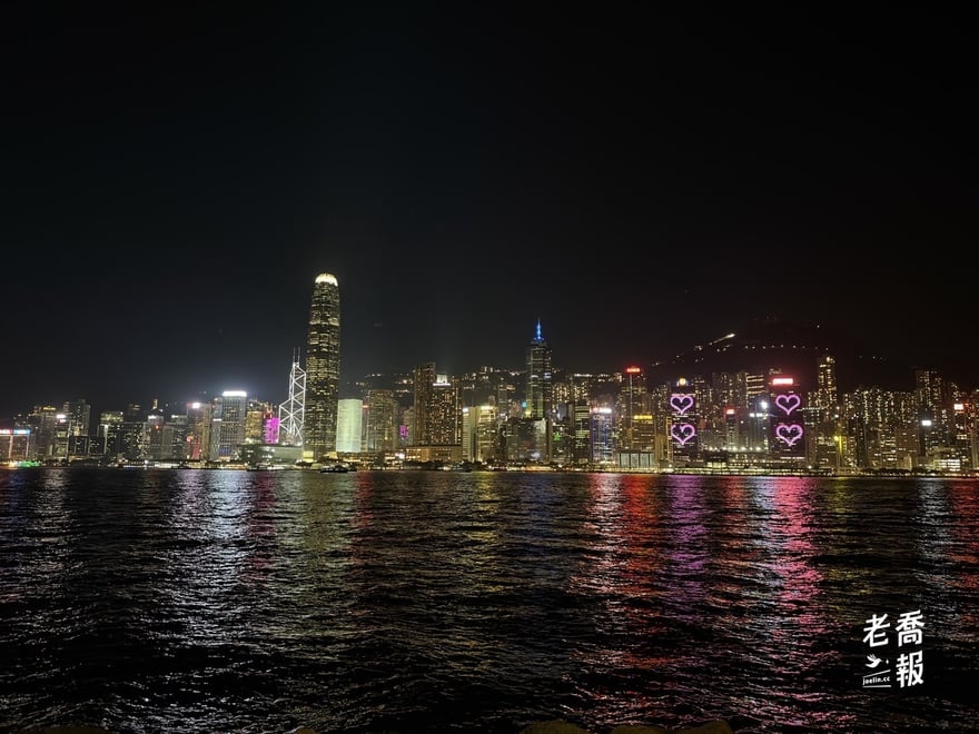 從九龍這側看維多利亞港夜景：對岸國際金融中心二期與中環的大樓燈火通明，幾棟大樓外牆亮著粉紅色愛心，倒影映在海面