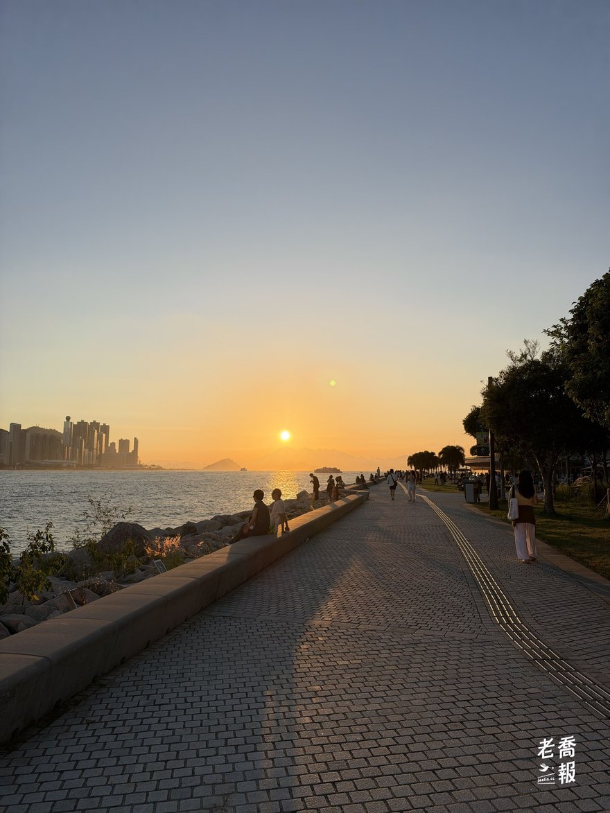 西九文化區海濱長廊的日落：太陽落在維多利亞港西邊的山後，海堤上有人散步、有人坐著看海