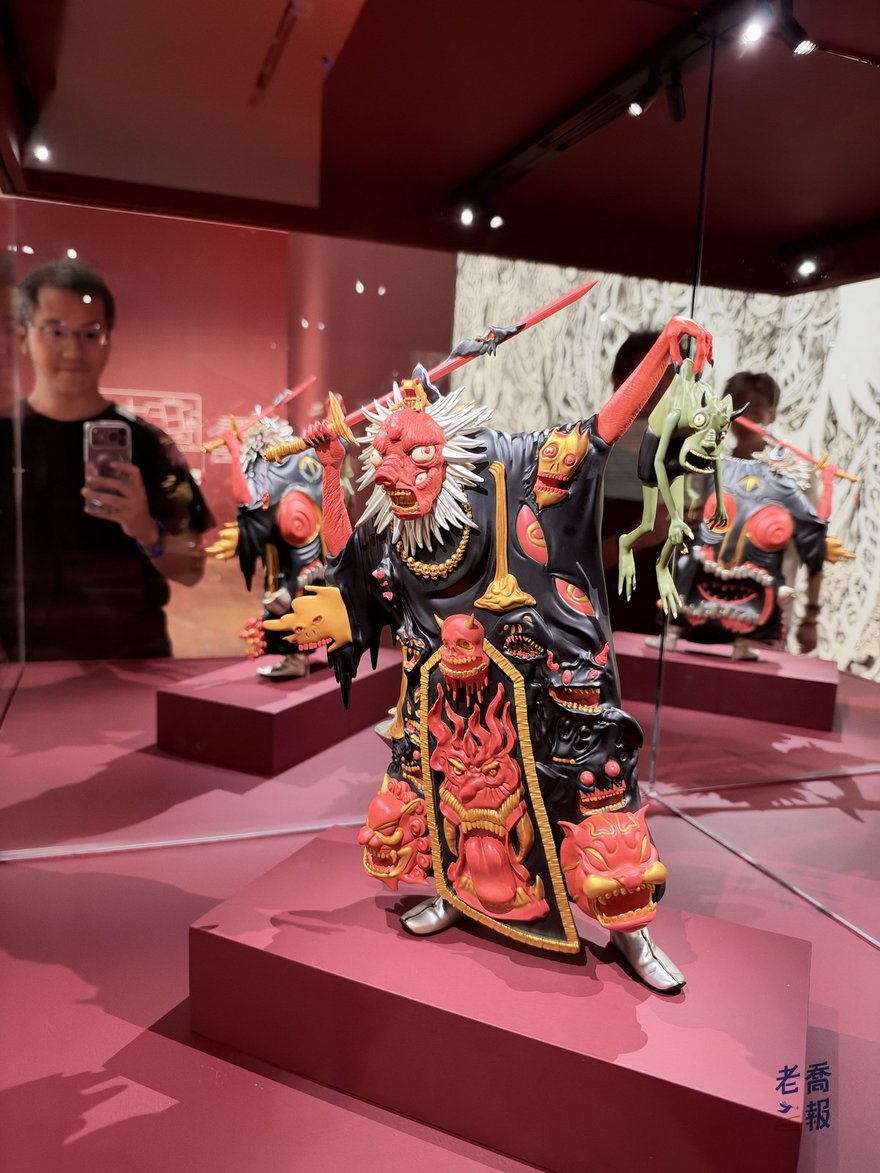 陳翊朗的大型公仔作品《鬼王》：紅臉白髮、一手高舉長劍、一手拎著綠色小鬼，黑色長袍上滿是紅色鬼臉，鏡面反射裡可以看到老喬在拍照