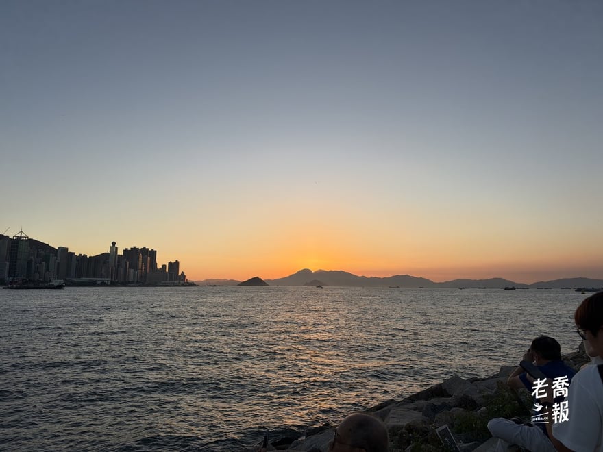 夕陽落在維多利亞港西邊的群山後方，左邊是港島西區的高樓剪影，岸邊的石堤上坐著看海的人