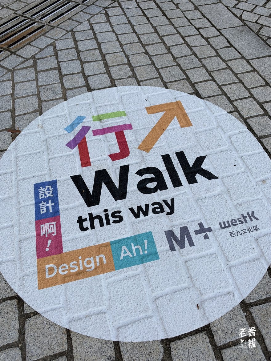 西九海濱步道地上的圓形指引貼紙，寫著「行 Walk this way」與「設計啊！Design Ah!」，下方是 M+ 和西九文化區標誌