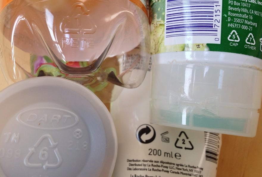 幾件塑膠製品放在一起：寶特瓶底壓印箭頭三角形 1 號與 PETE、白色杯蓋壓印 6 號、乳液瓶背標印著三角形 2 號，另一個瓶身標籤印著 5 號 CAP 與 7 號 OTHER。