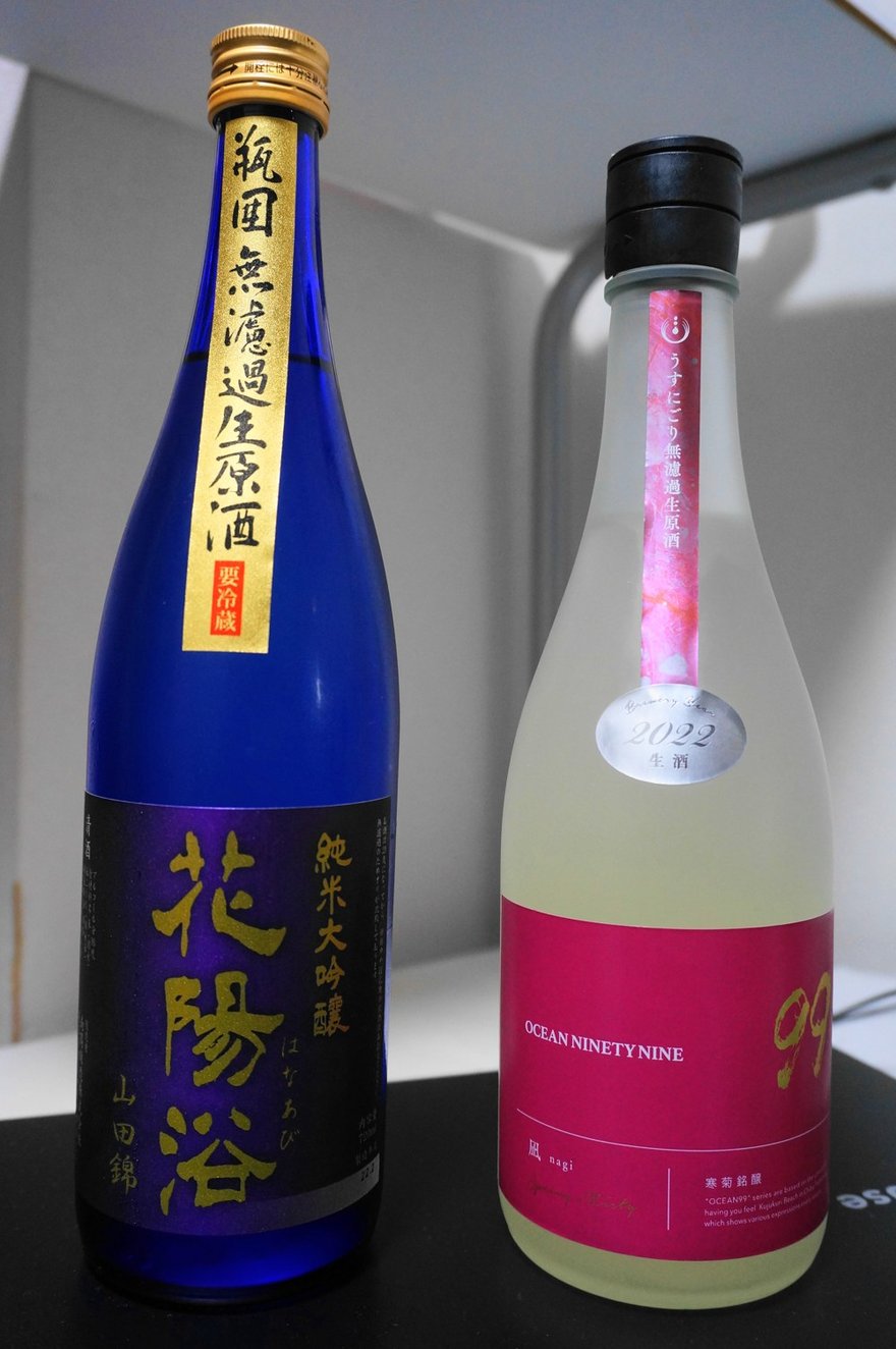 兩支日本酒瓶，左邊藍色瓶的酒標上寫著「純米大吟醸」「山田錦」，瓶頸的黃色肩標寫著「無濾過生原酒」「要冷蔵」。