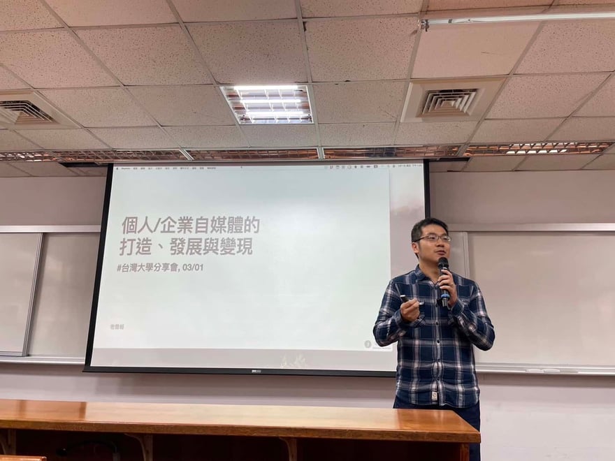 教室裡講者手持麥克風站在投影幕前，投影片標題為「個人／企業自媒體的打造、發展與變現」，副標為台灣大學分享會 03/01