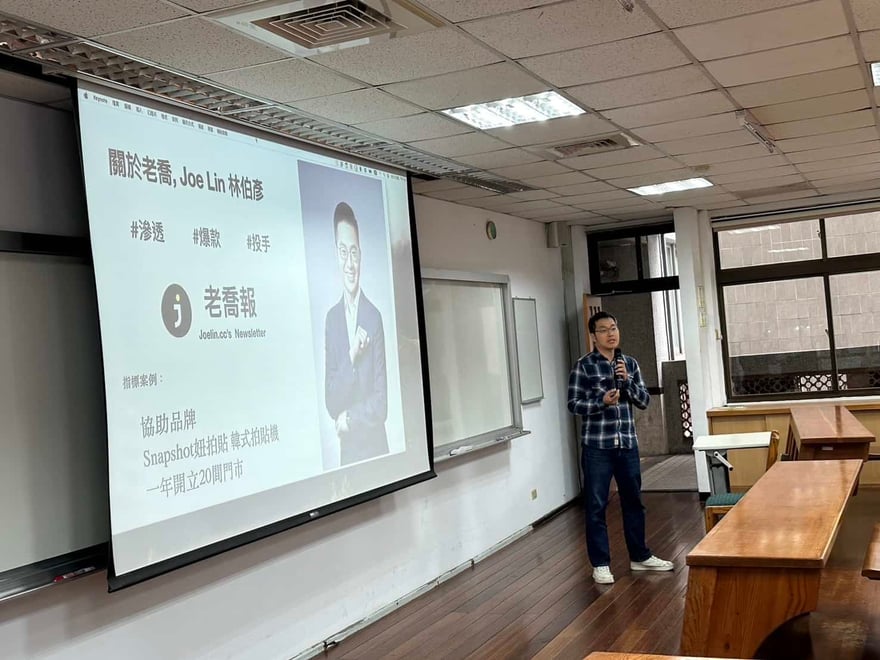 教室內講者拿著麥克風站在投影幕旁，投影片為自我介紹頁，提到老喬報與協助 Snapshot 妞拍貼韓式拍貼機一年開立 20 間門市
