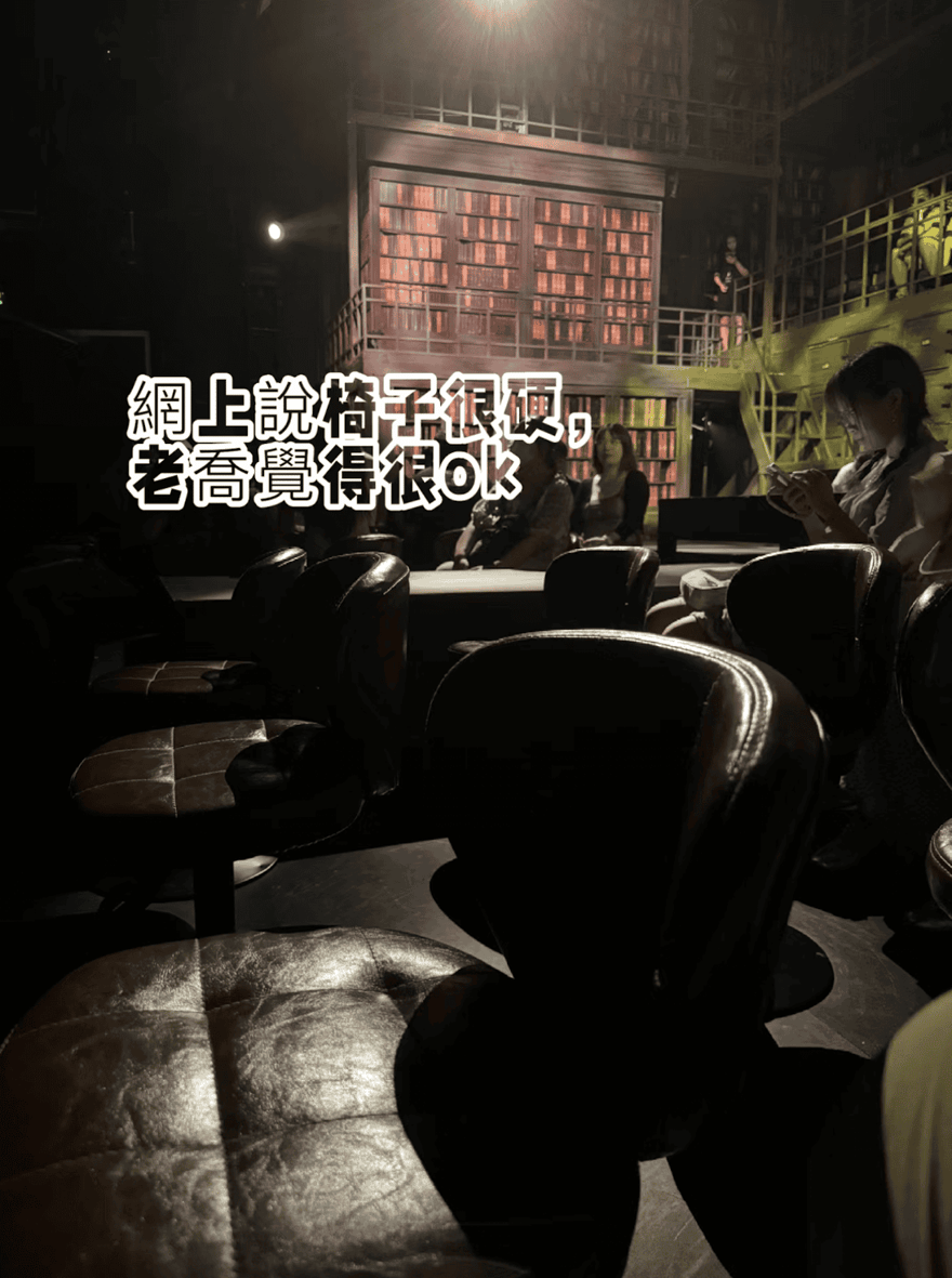 昏暗劇場中的皮革旋轉座椅，遠處是紅褐色書架佈景與觀眾，圖上字幕寫網上說椅子很硬，老喬覺得很 ok