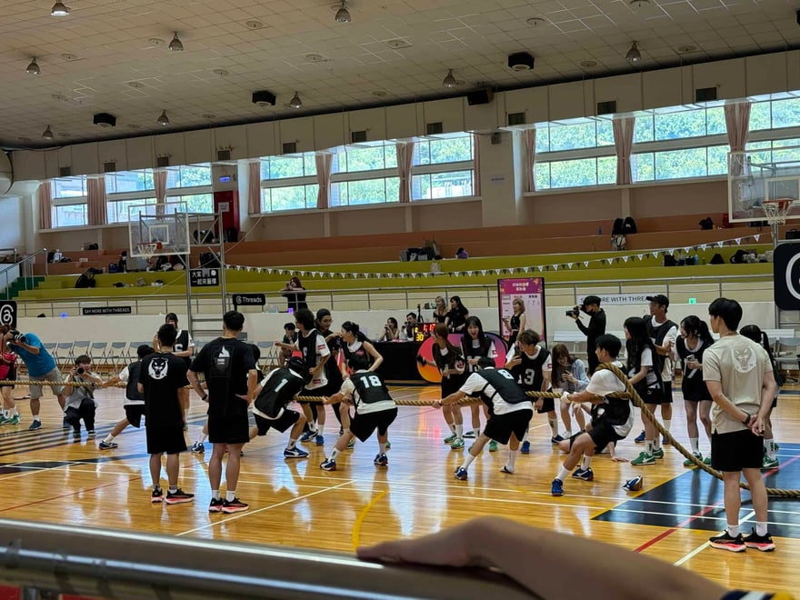 室內體育館木地板上，兩隊穿黑白運動服的人正在進行拔河比賽，場邊有人拍攝與觀賽，看台與牆邊掛著 Threads 看板