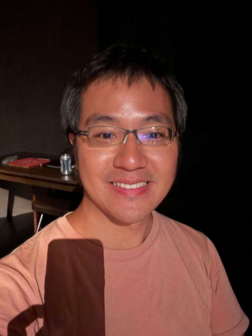一位戴眼鏡、穿粉橘色 T 恤的男子在昏暗的劇場內自拍，背後可見木桌與桌上的啤酒罐和紅色筆記本