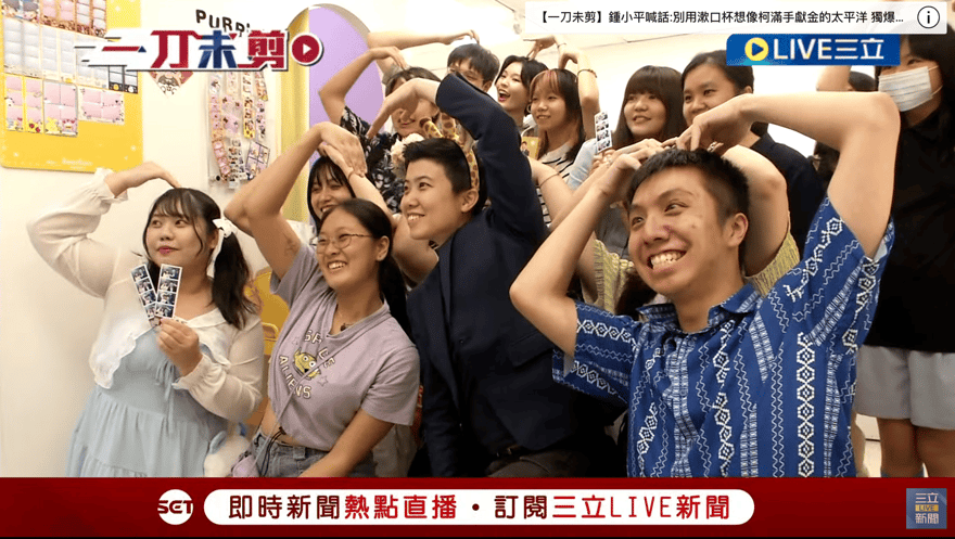 三立新聞直播畫面截圖，一群人在拍貼店內雙手在頭上比出愛心合影，中間穿深色西裝的是苗博雅，左側女生手拿兩條拍好的拍貼