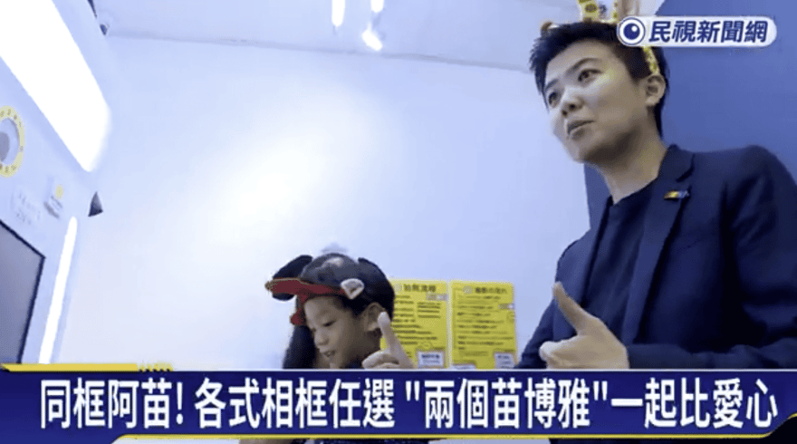 民視新聞網畫面截圖，穿深色西裝、頭戴黃色動物髮箍的人與戴黑紅髮箍的小孩在拍貼機前比手勢，字幕寫同框阿苗！各式相框任選兩個苗博雅一起比愛心