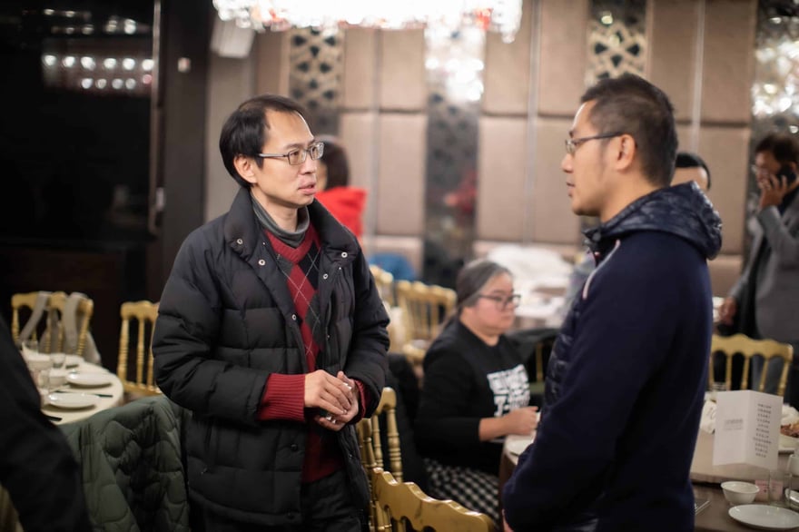 宴會廳內兩位戴眼鏡的男士面對面交談，左邊穿黑色羽絨外套，右邊穿深藍外套，背景是圓桌與金色椅子。