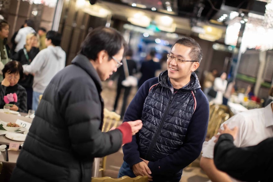 宴會現場，一位穿深藍背心外套的男士微笑聽著穿黑色羽絨外套的男士說話，背景有賓客走動。
