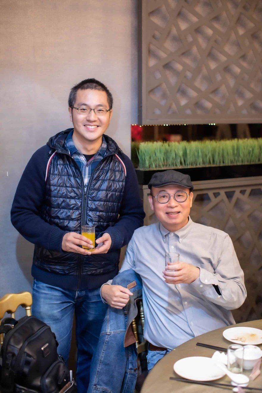 兩位男士在宴會廳合影，站著的穿深藍背心外套、微笑拿著果汁，坐著的戴貝雷帽、穿灰藍襯衫，兩人都拿著玻璃杯。