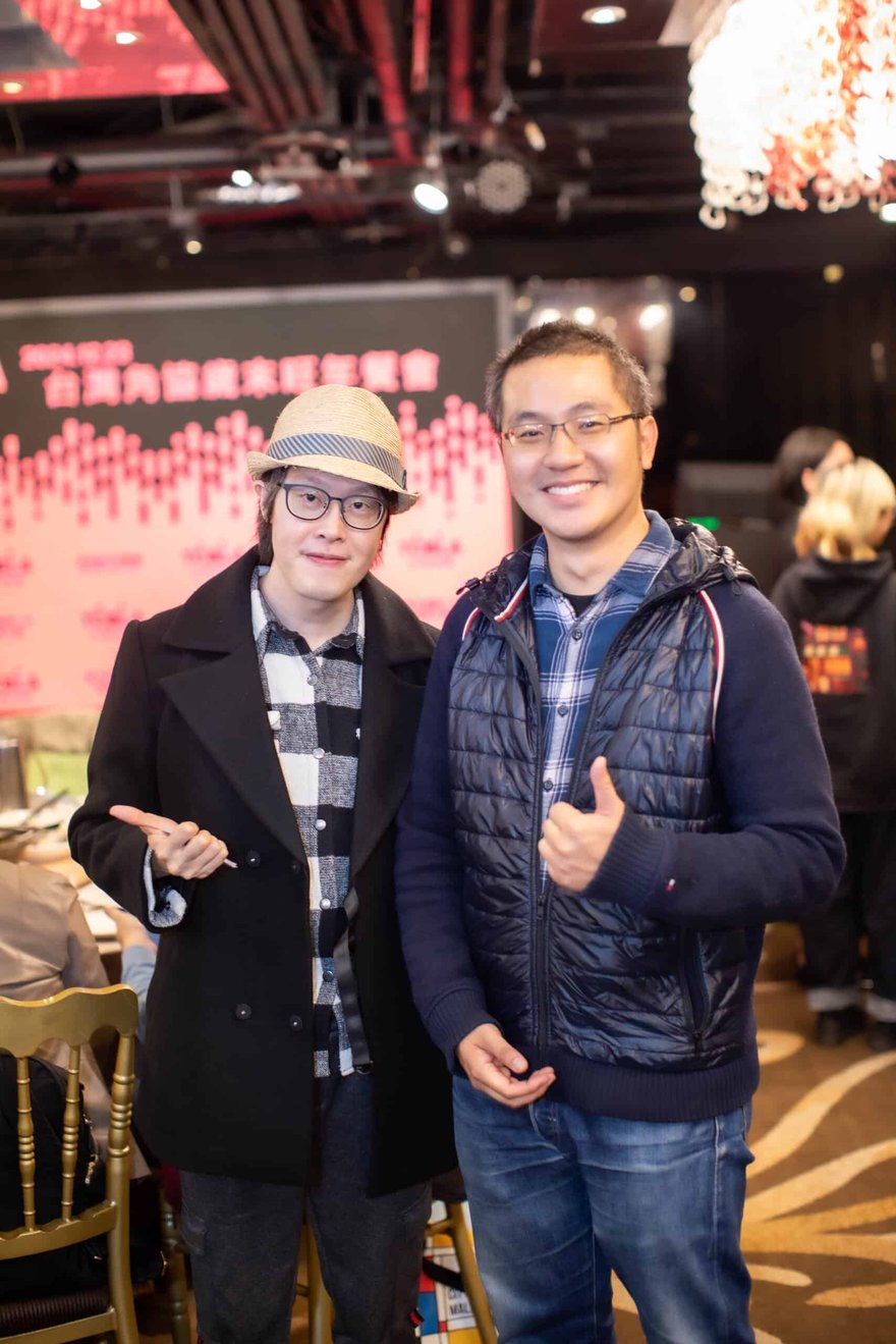 兩位男士在宴會現場合影，左邊戴草帽、穿黑外套，右邊穿深藍背心外套並比讚，背後是活動背板。