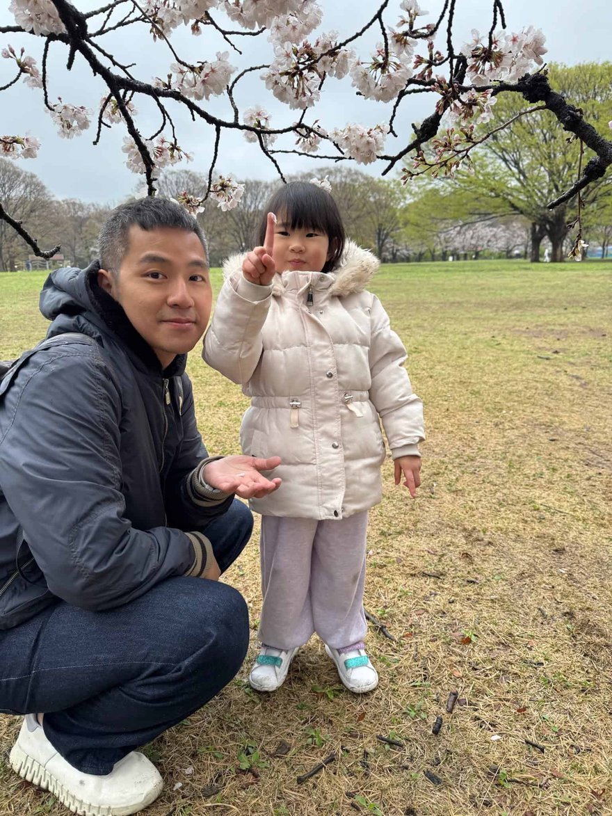 大人蹲在盛開的櫻花樹枝下，旁邊穿米白色羽絨外套的小女孩比出手勢面對鏡頭
