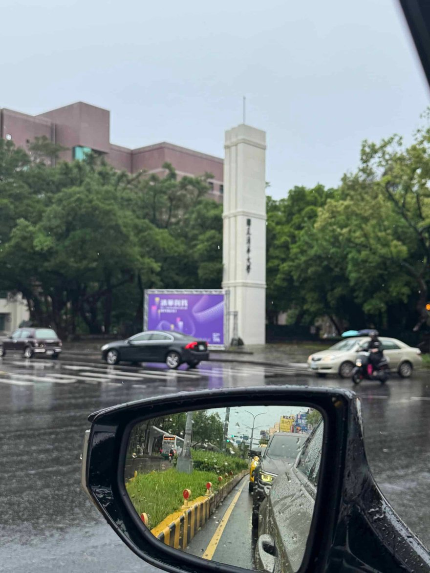 雨天從車內拍到的清華大學校門白色柱狀入口，旁邊立著紫色看板，後照鏡裡可見路上的車輛