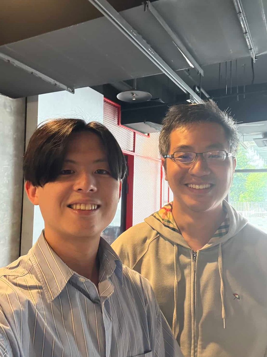 兩位男士在室內自拍合照，左邊是穿藍白條紋襯衫的年輕男子，右邊是戴眼鏡穿灰色連帽外套的男子，兩人都面帶笑容