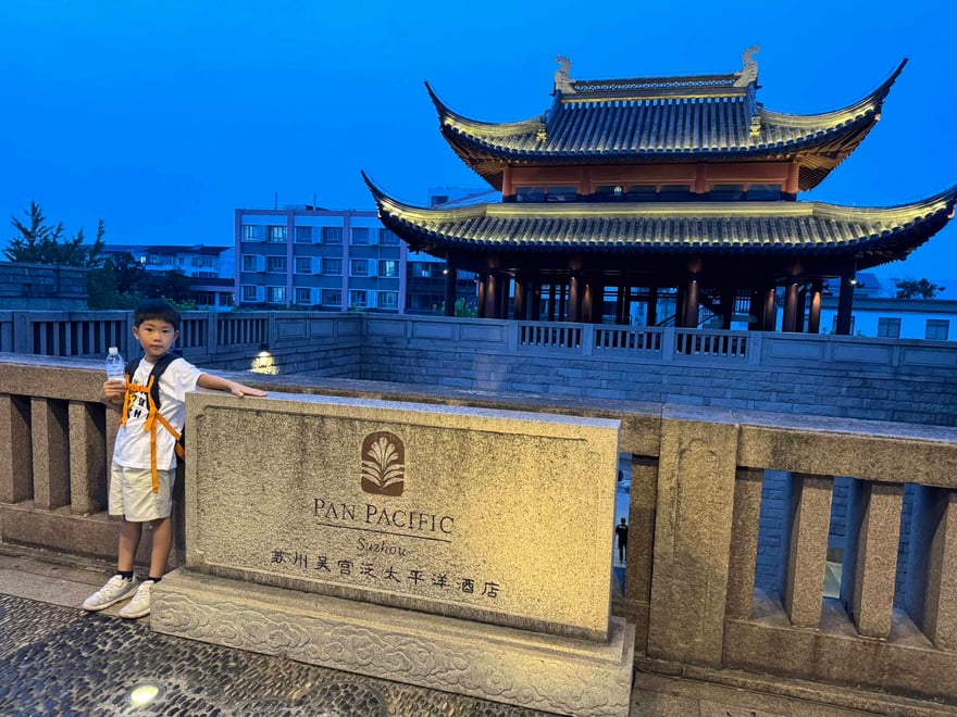 Traditional Chinese pavilion with ornate roof design at dusk, featuring a young boy holding a drink in front of a stone sign for Pan Pacific hotel, showcasing cultural architecture and urban scenery.