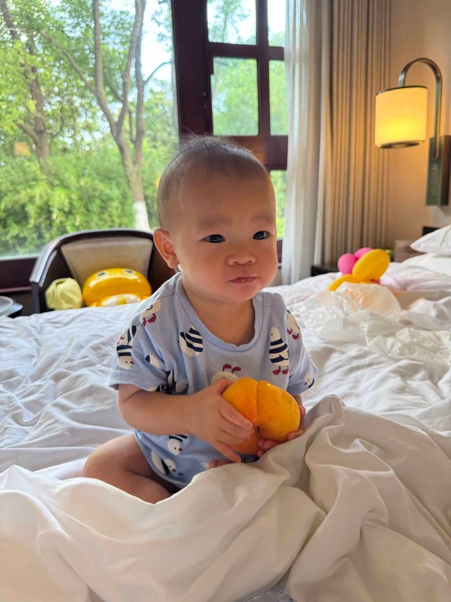 Adorable baby sitting on a bed holding a yellow fruit, with a bright window and cozy interior background, perfect for family and parenting content.