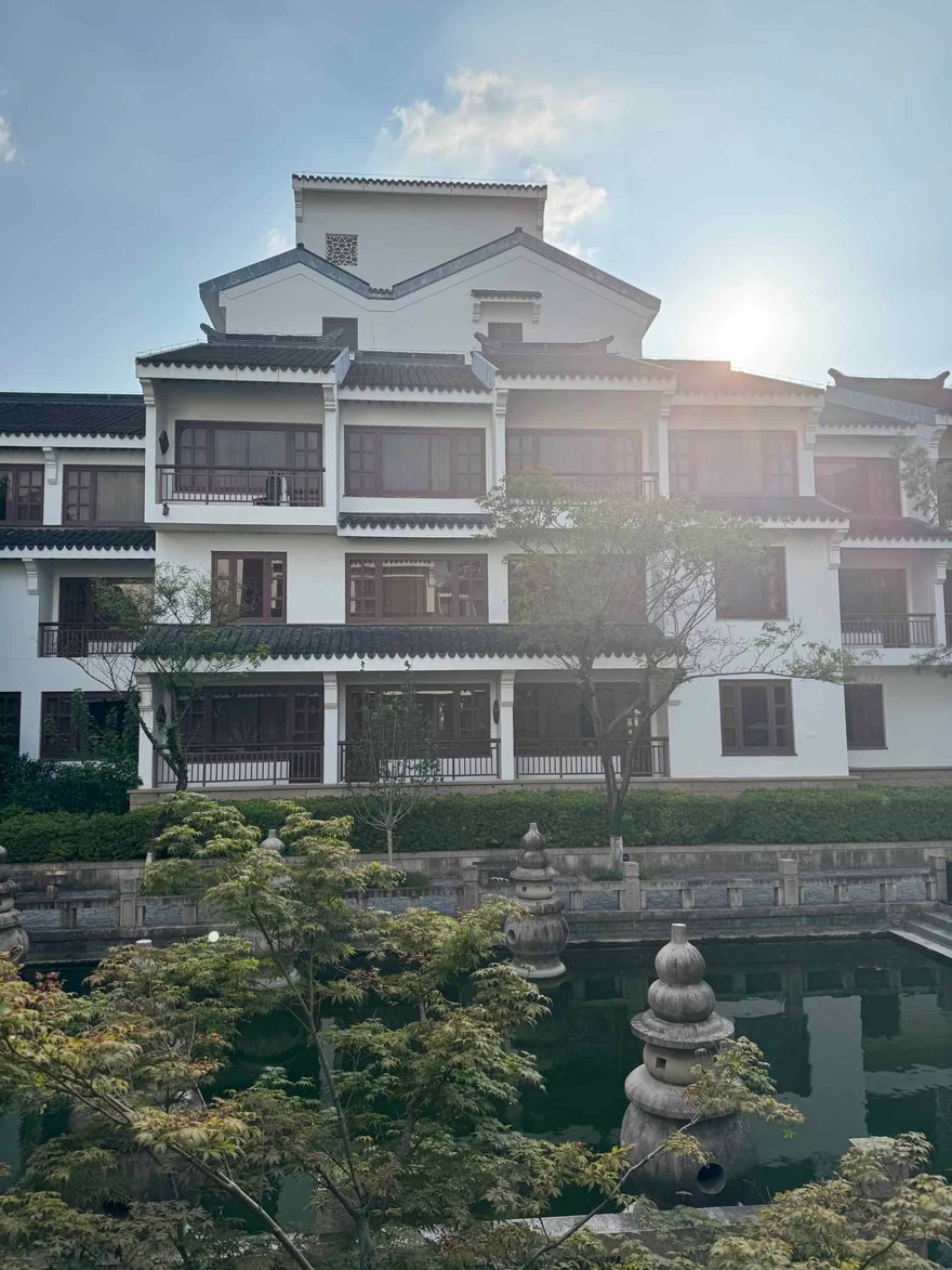 Style building with white walls, dark wooden accents, and tiled roofs, situated by a serene pond with lush greenery and stone lanterns, under a bright sky.