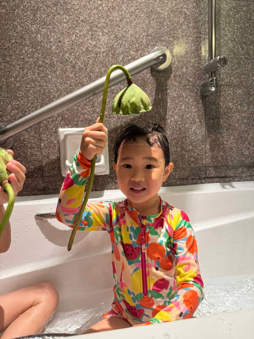 A colorful young girl enjoying a bath in a public bathhouse, holding a green lotus flower-shaped bath sponge, with a cheerful expression and vibrant floral-patterned swimsuit.
