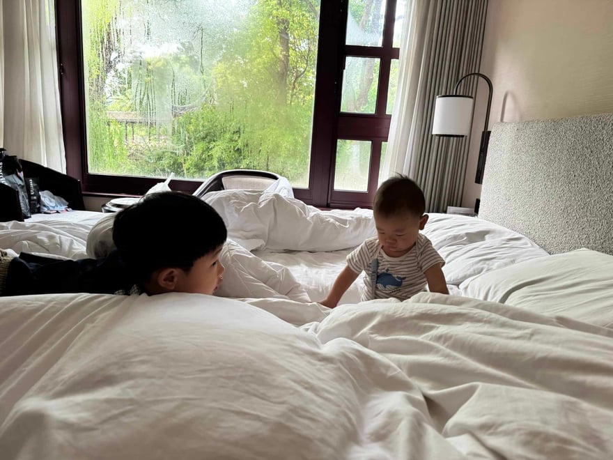 Two young children playing and interacting on a large, cozy hotel bed with white linens, near a window with lush greenery outside, in a bright, modern hotel room setting.