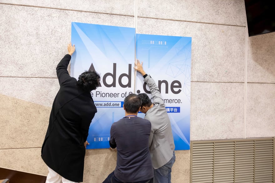 Add.one logo being displayed at a conference or event, with three individuals installing or unveiling the sign on a wall. The scene emphasizes technology, innovation, and corporate branding.