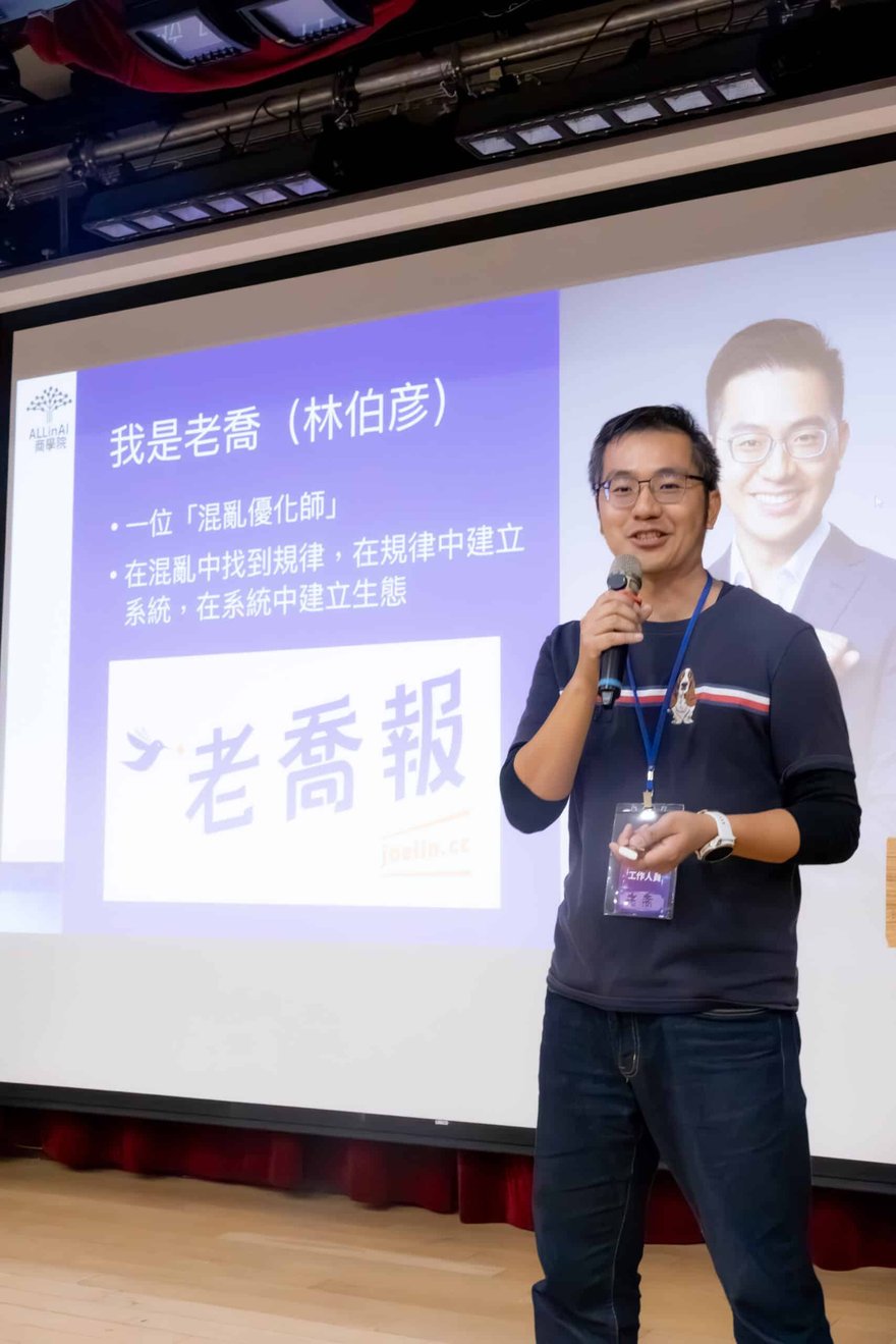 老喬在台上拿著麥克風演講，身後投影片寫著「我是老喬（林伯彥）」與「混亂優化師」，並有老喬報標誌，他胸前掛著工作人員證。