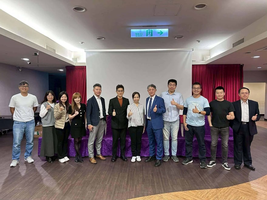 活動現場的講師合影，十二位講師與工作人員站在紫色舞台前，多人比讚，後方是白色投影幕與紅色布幔。