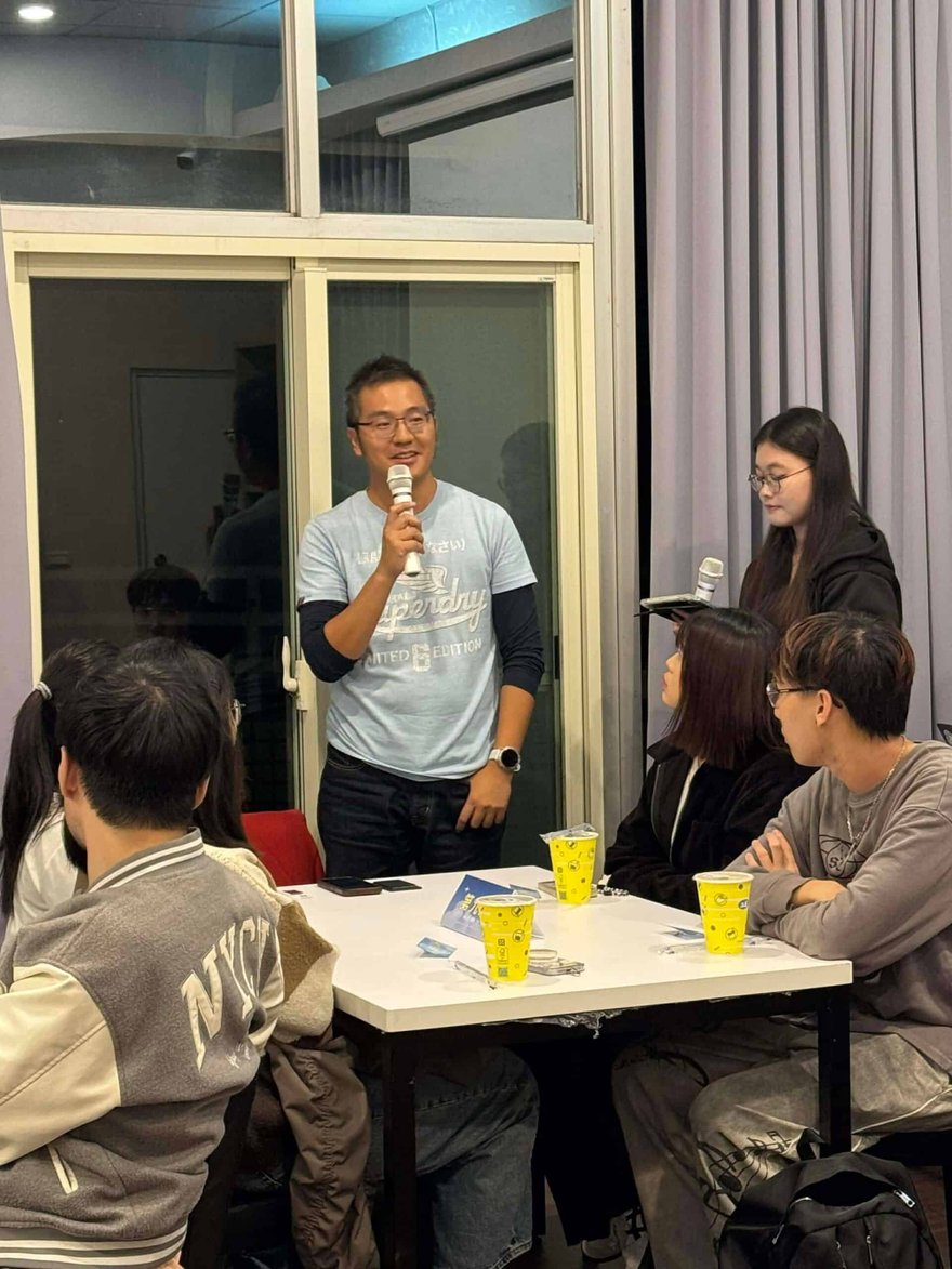 老喬在活動現場拿著麥克風分享，穿淺藍色上衣，面前是白色桌子與黃色飲料杯，周圍坐著聆聽的學生。
