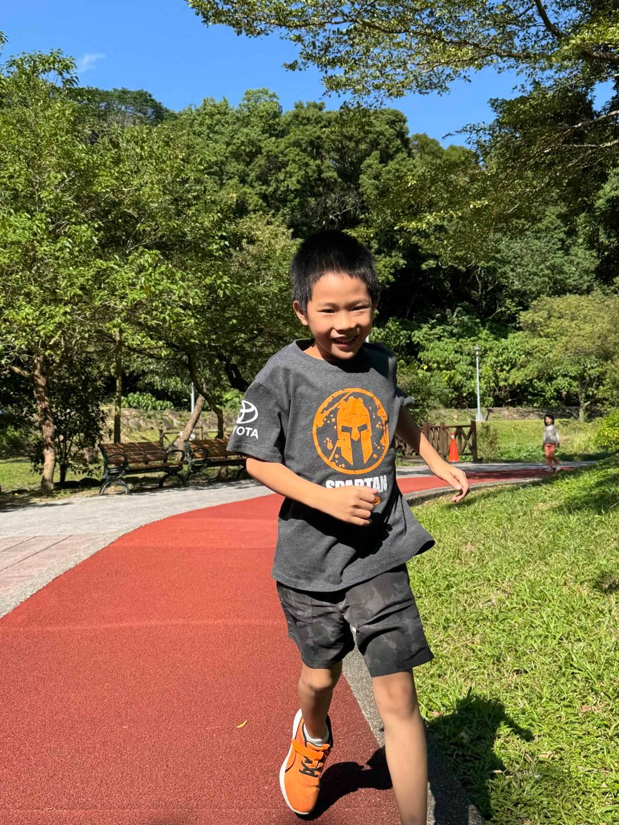 Running child in a park with lush green trees, wearing a gray T-shirt with an orange logo, black shorts, and orange sneakers, enjoying outdoor activity on a sunny day.