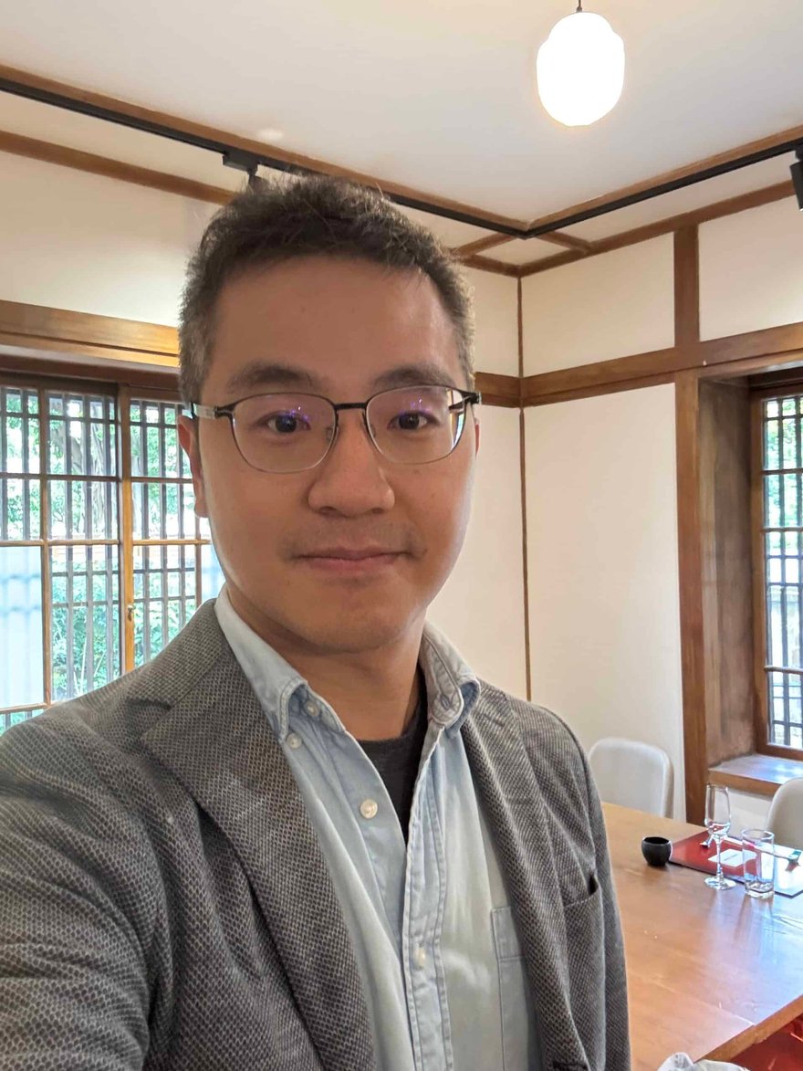 A. ALT tag: Professional portrait of a man in a modern office setting, wearing glasses and a blazer, with natural light and wooden interior design elements.
