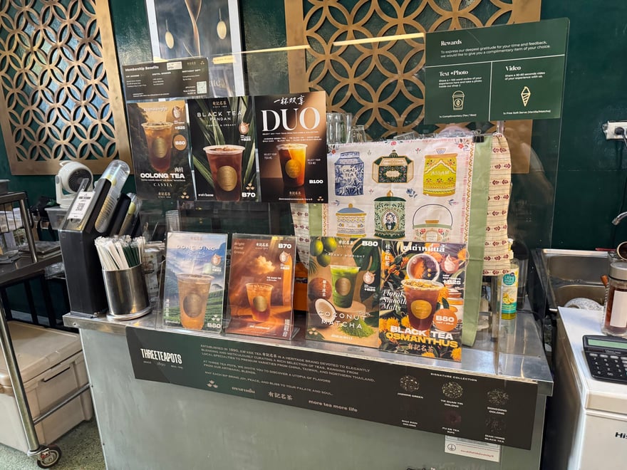 THREE TEA POTS 店內吧台，立著多款飲品菜單與價目的牌卡，牆面是綠色底配幾何花窗，標示茶飲 70 至 100 泰銖