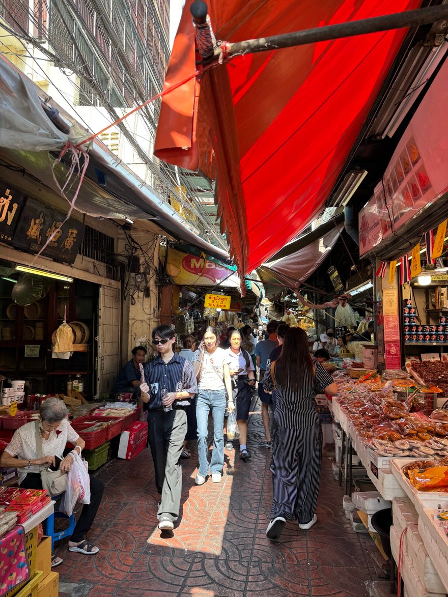 曼谷唐人街的市場巷弄，兩側店家與攤位擠在紅色遮陽棚下，行人走在紅磚地上