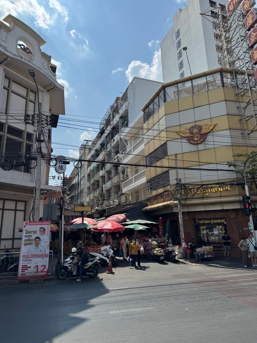 曼谷唐人街的街口，陽光下可見黃色大樓與翅膀標誌，路邊有遮陽傘攤位與摩托車
