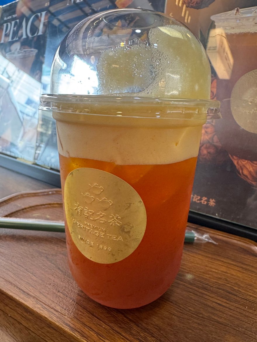 一杯上層有奶蓋的橘色冰茶，杯身貼著有記名茶 Heritage Tea 的金色圓形貼紙，放在木盤上