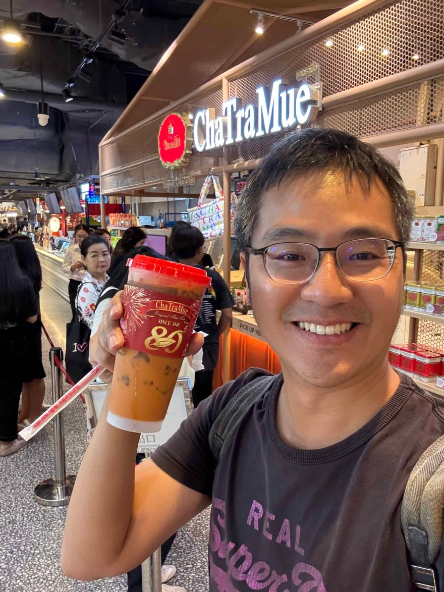 男子在 ChaTraMue 店前舉著一杯紅色杯身的泰奶自拍，身後是排隊人群與店面